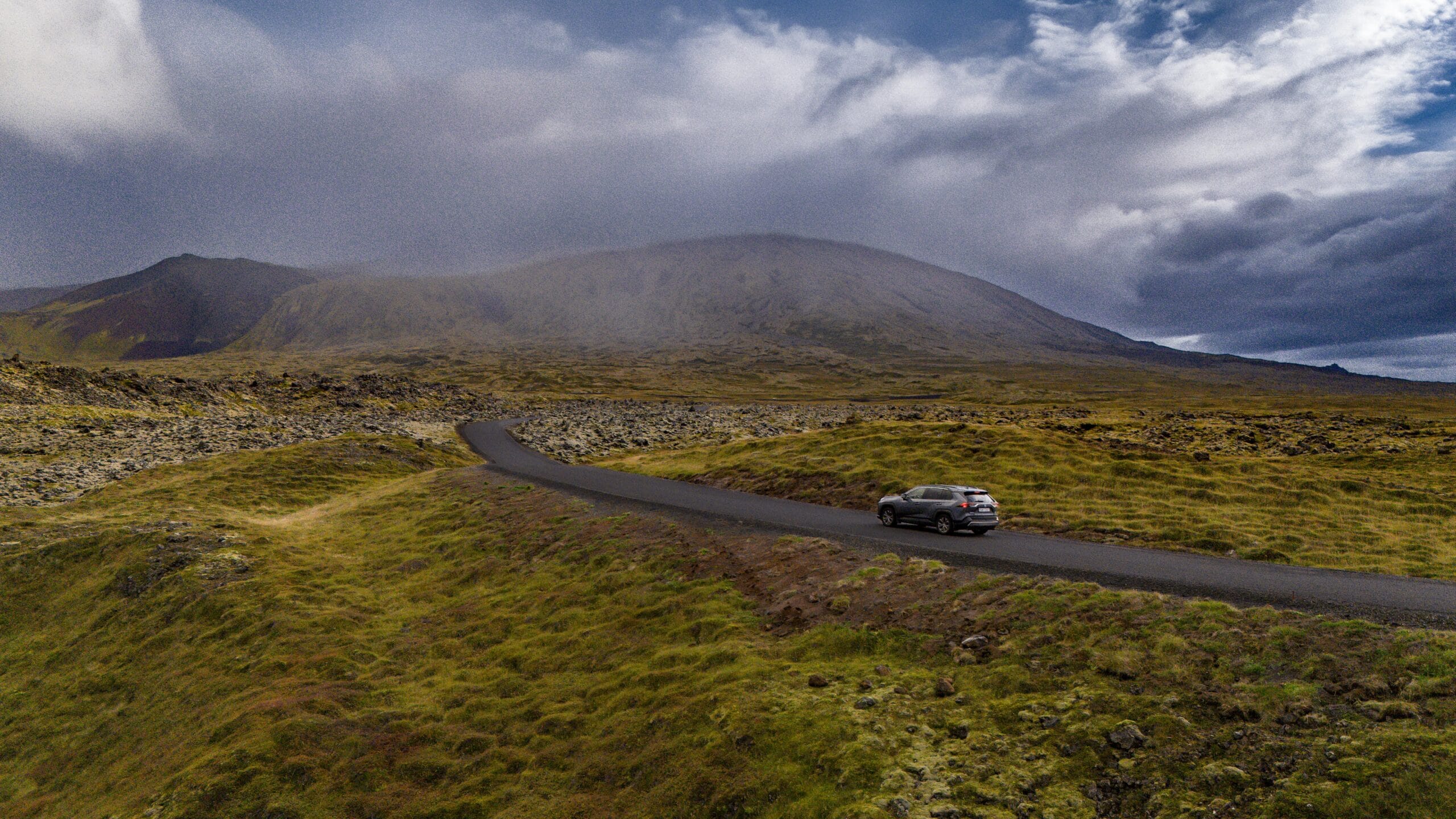 A lone car travels on a winding road through a vast, rugged landscape under a dramatic, cloudy sky. Surrounding hills and sparse vegetation evoke a sense of solitude.