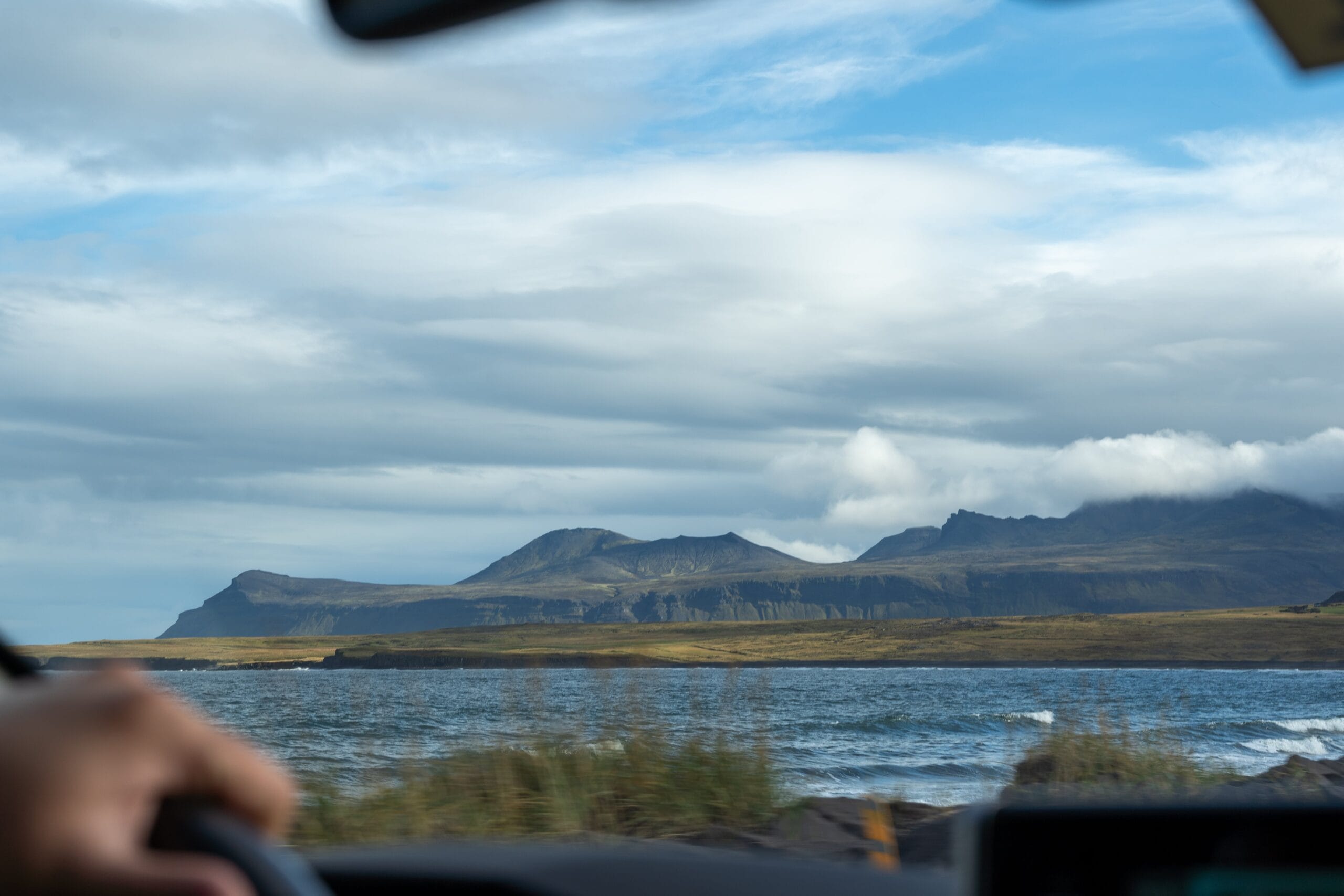 View from inside a car showing a serene landscape with a calm lake, grassy shore, and distant mountains under a partly cloudy blue sky.