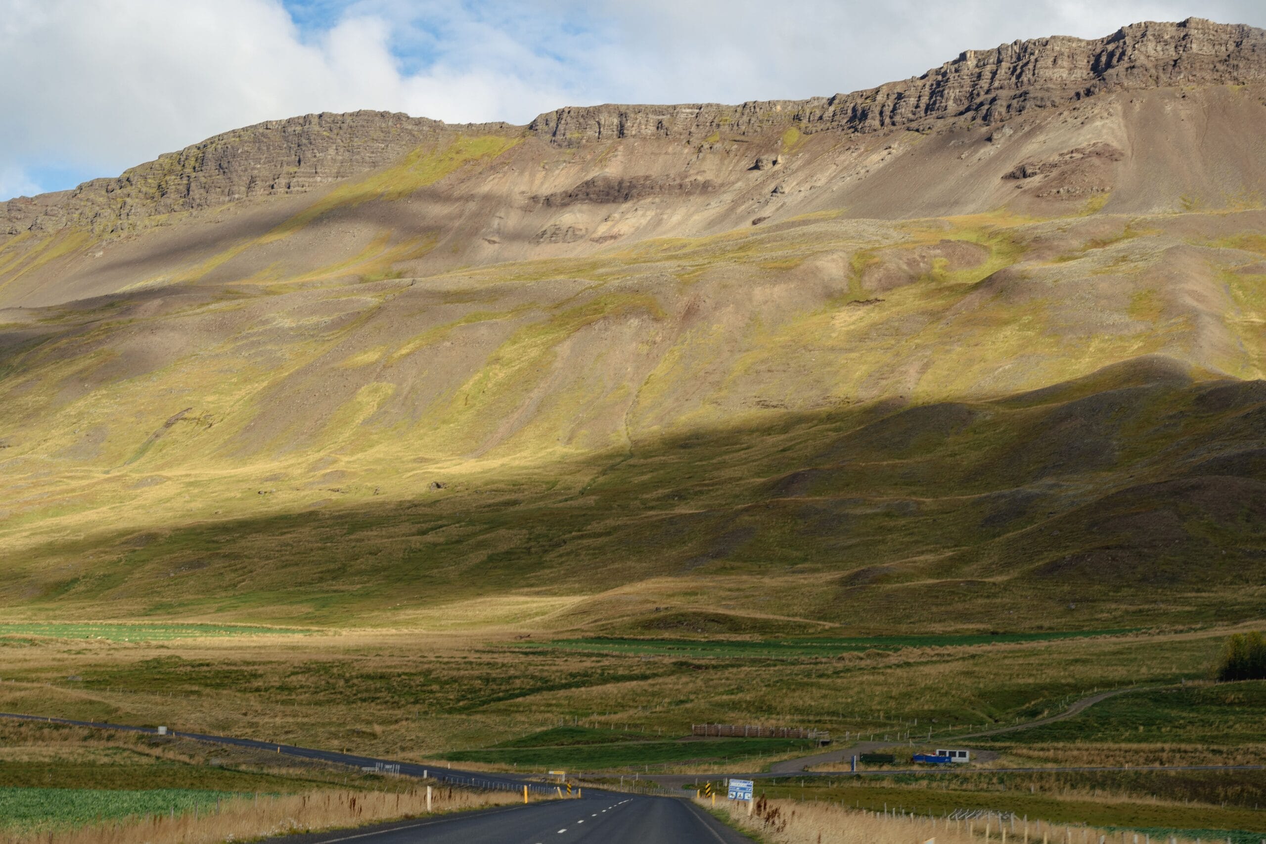 A winding road leads towards a rugged mountain range under a partly cloudy sky. Sunlight casts dynamic shadows on the grassy terrain, creating a serene mood.