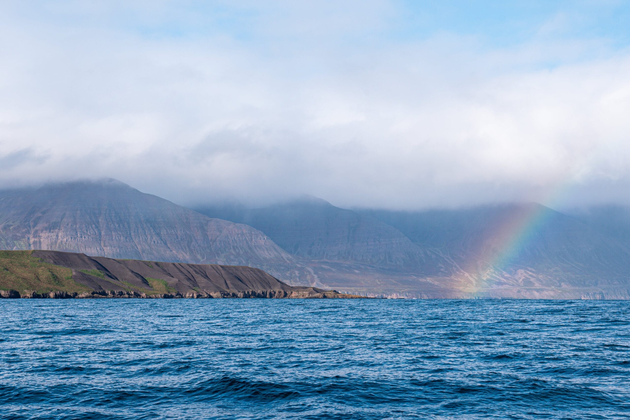 Ocean meets a mountainous shoreline under cloudy skies, with a vivid rainbow arching over the hills, creating a serene and uplifting scene.