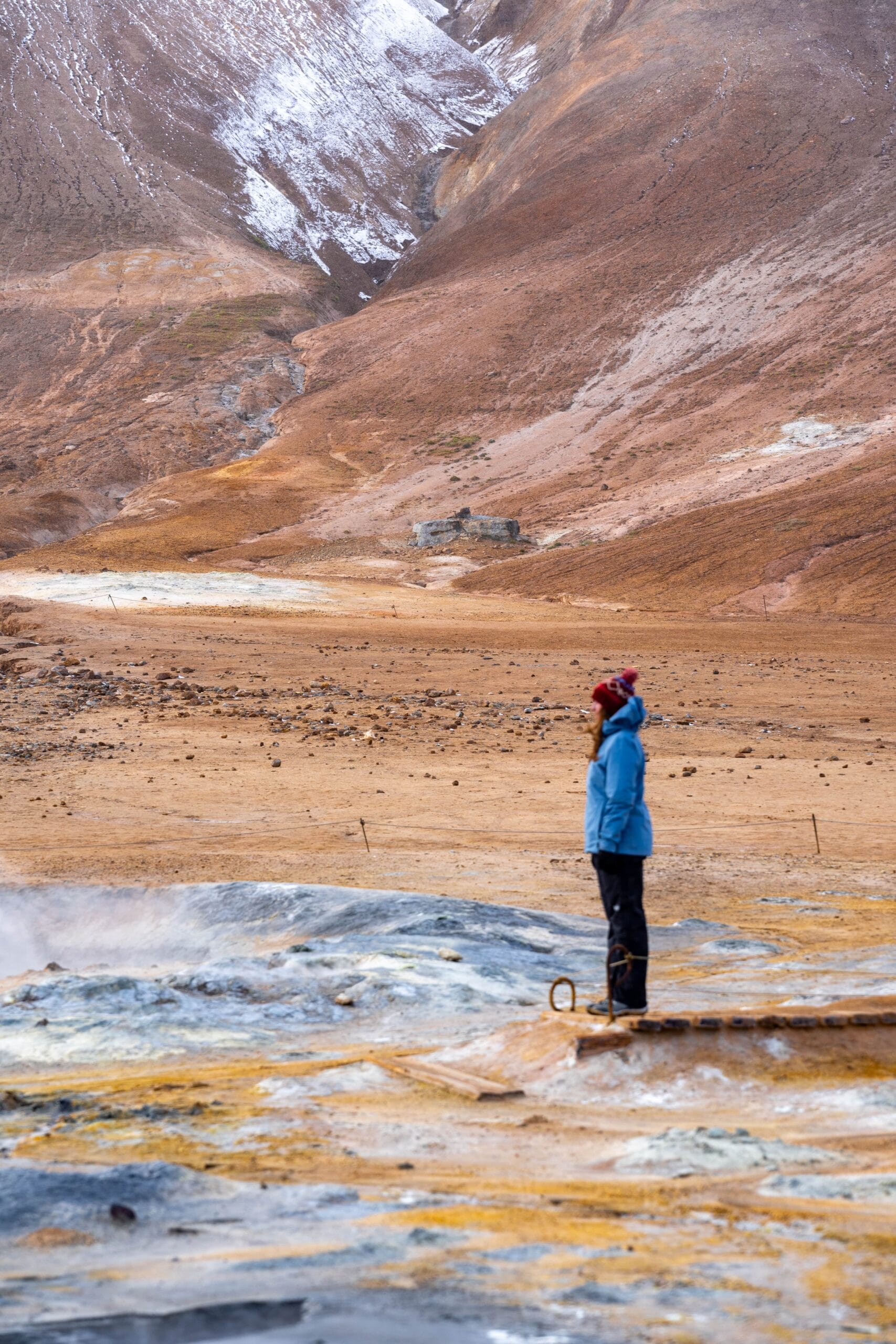 A person in a blue jacket and red hat stands next to a steaming geothermal area, set against a vast, rugged landscape with brown hills and patches of snow, conveying a sense of solitude and awe.