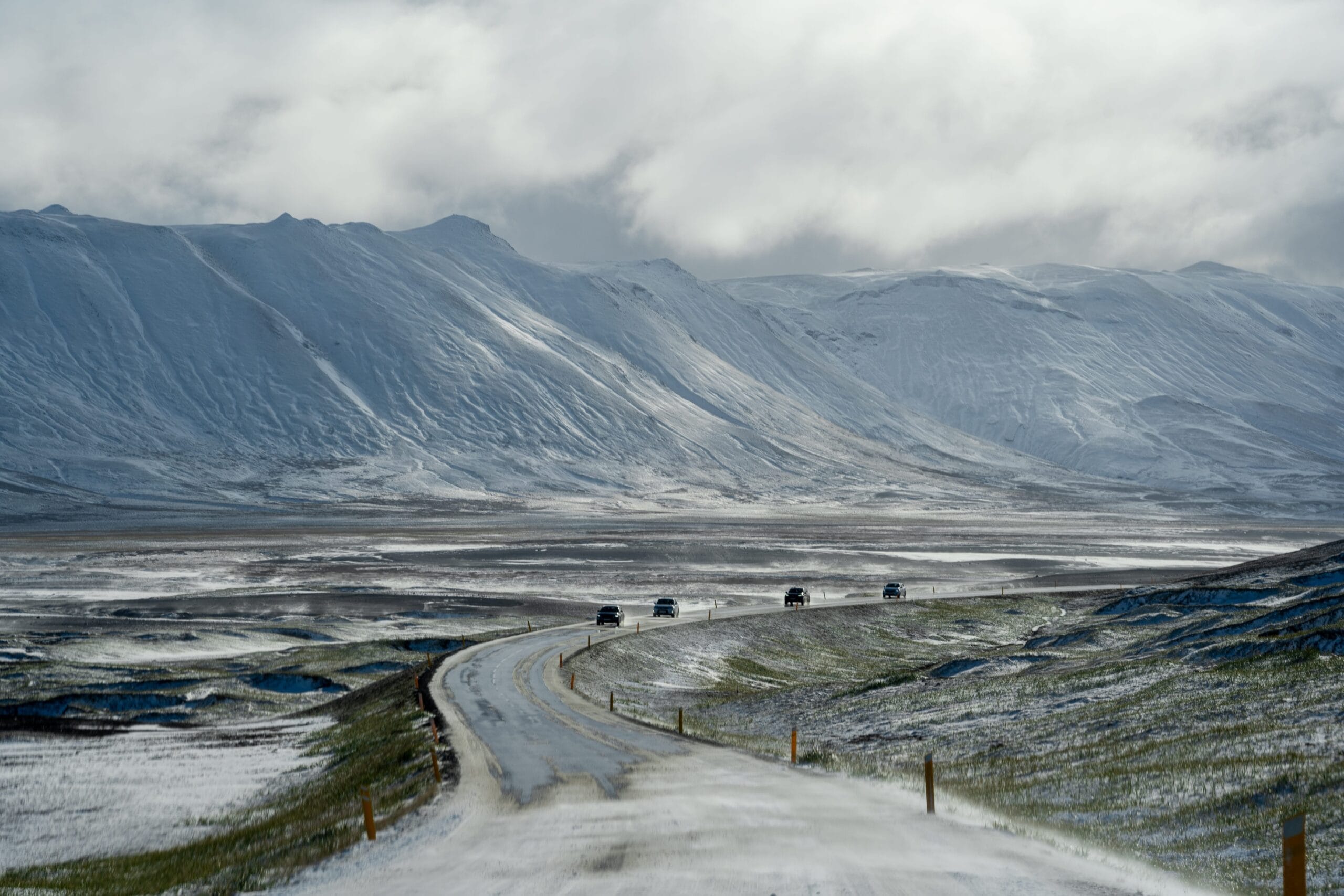 A winding road covered in light snow leads towards distant, snow-capped mountains under a cloudy sky. Four cars drive along the road, conveying a sense of journey and adventure.