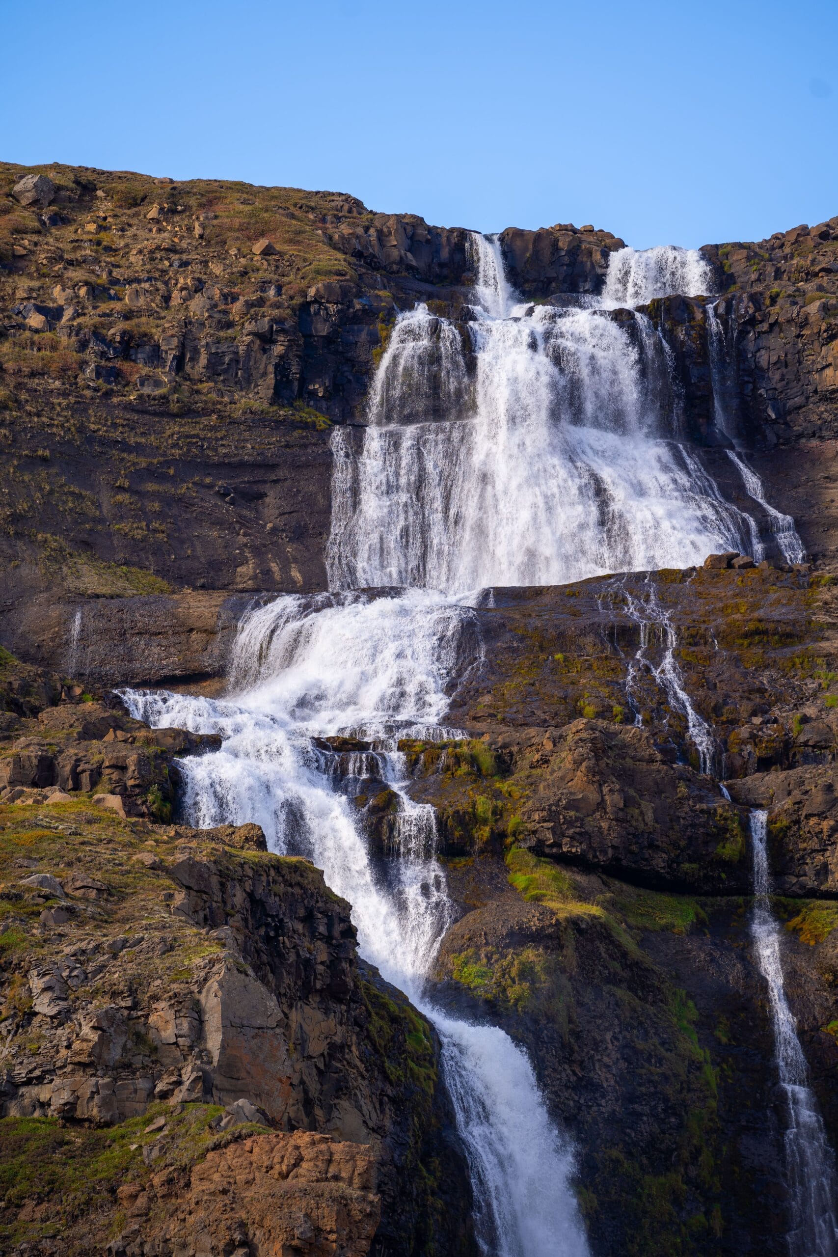 A majestic waterfall cascades over a rugged cliff, with multiple tiers spilling water onto rocks below. The scene is serene under a clear blue sky.