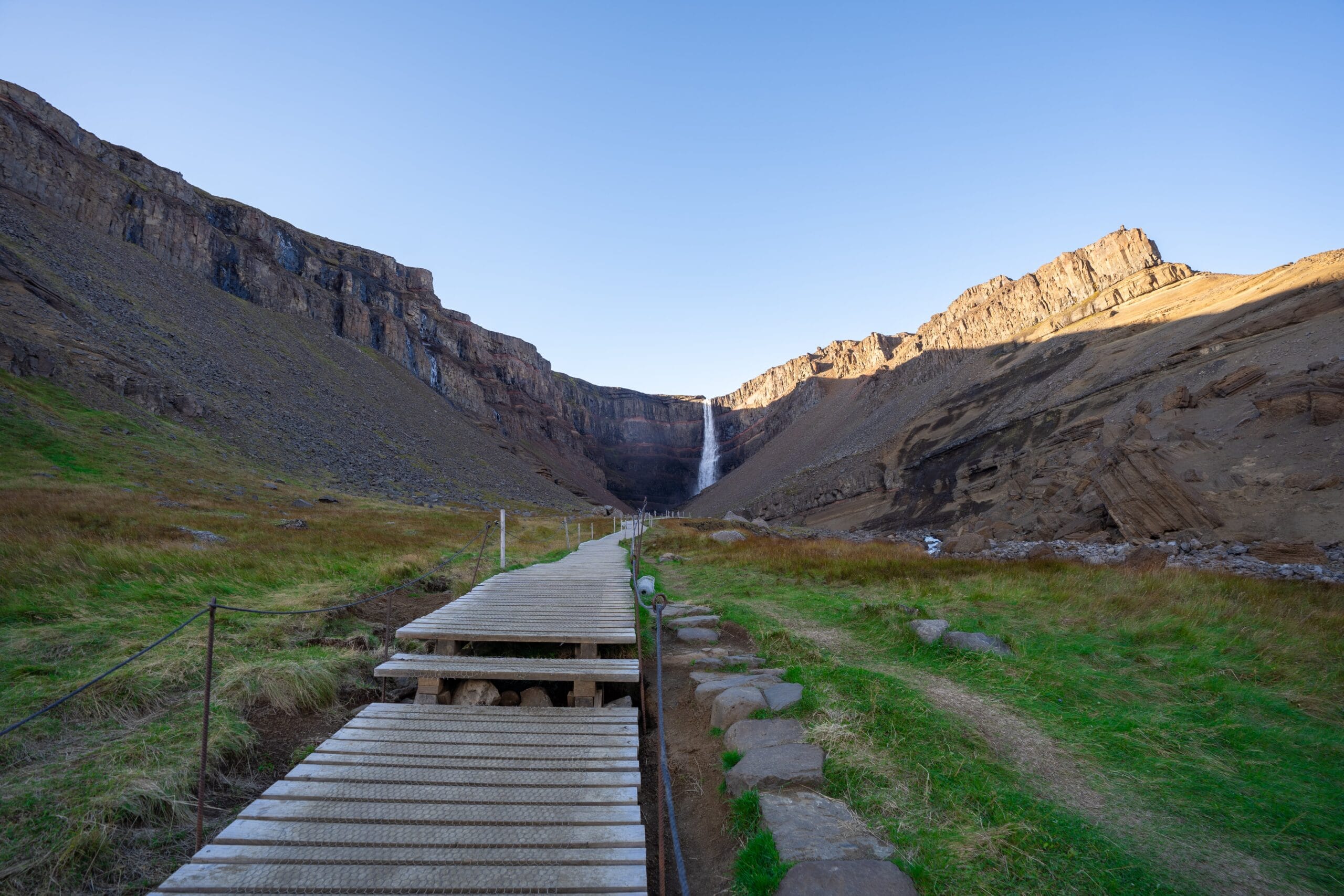 Wooden path leading to a distant waterfall, flanked by grassy areas and rocky cliffs under a clear blue sky, conveying a serene and inviting atmosphere.