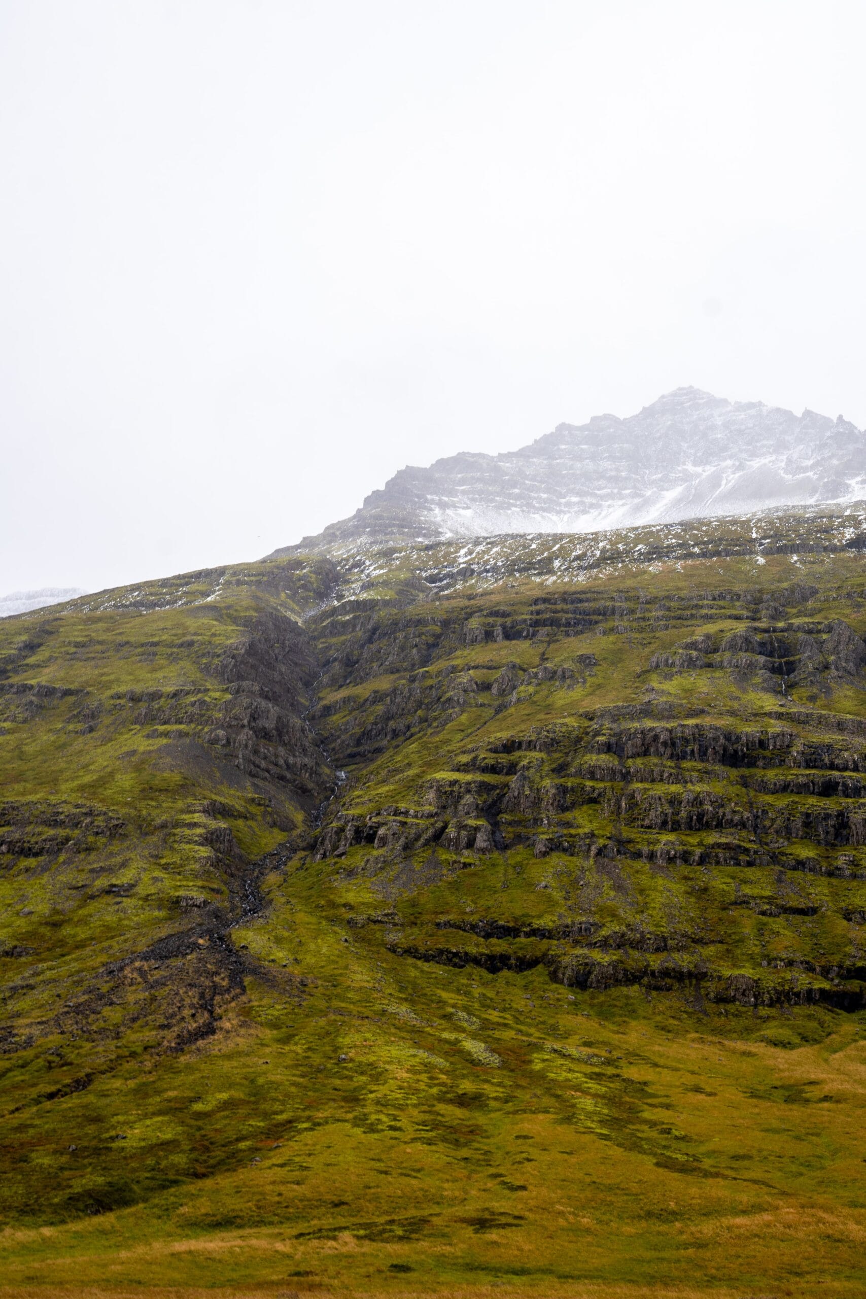 A serene mountain landscape features a grassy slope with dark rock layers, leading to a snow-dusted peak under an overcast sky, conveying solitude and calm.