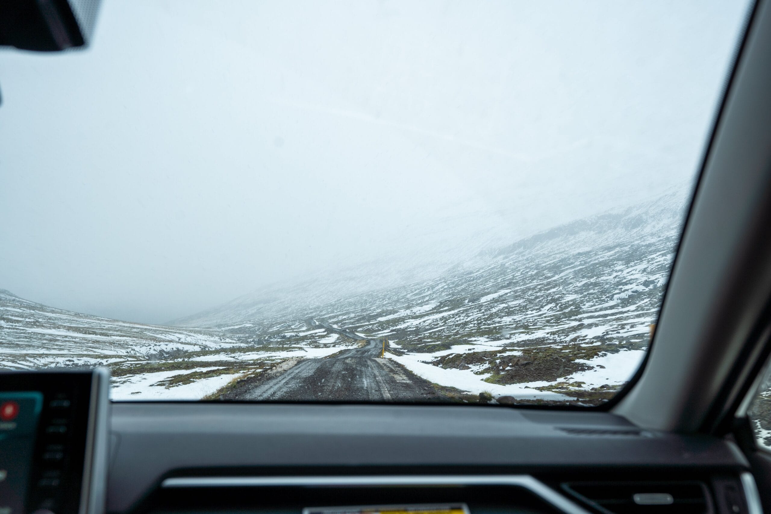 View from a car interior showing a narrow, snowy road winding through a desolate, foggy mountain landscape, conveying a sense of isolation and adventure.