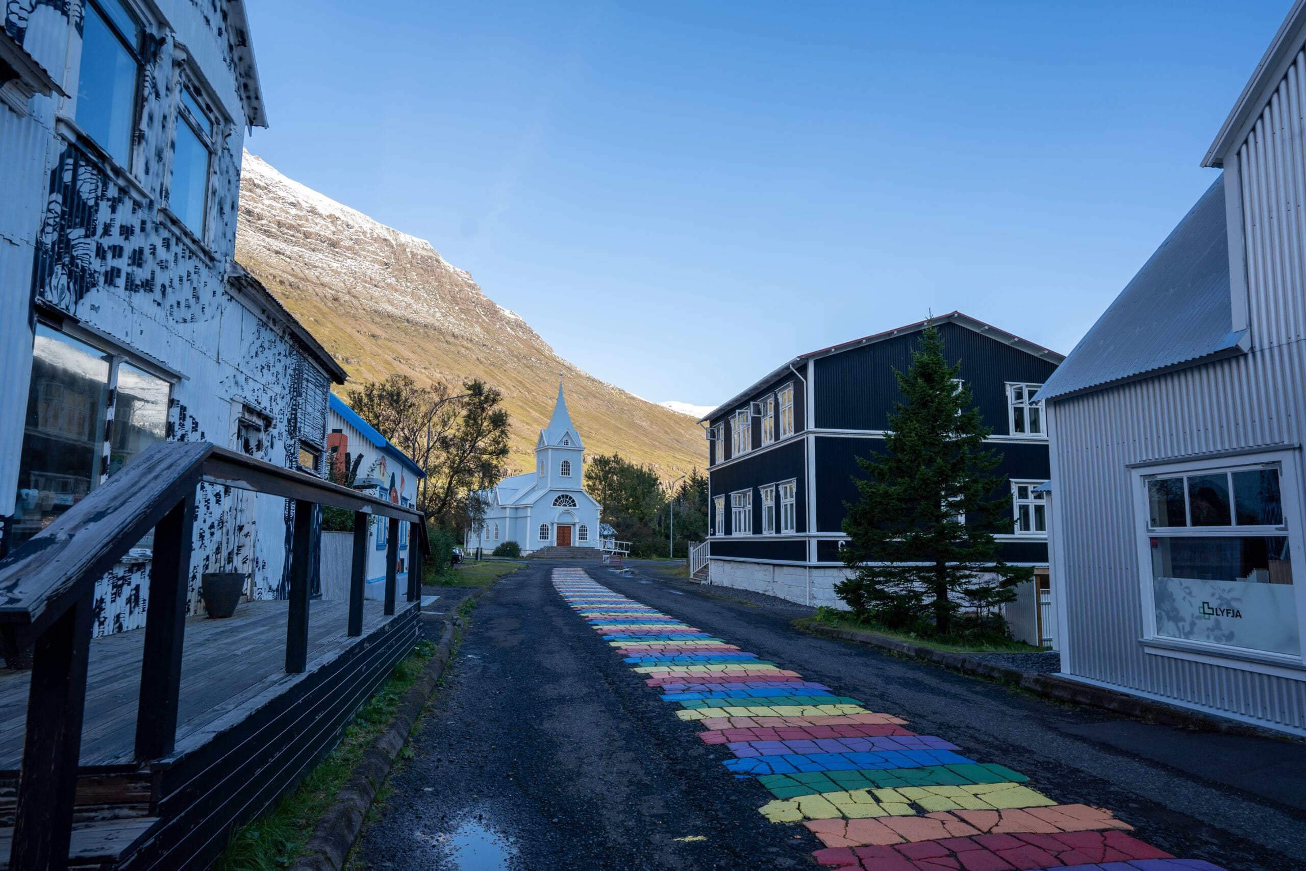 A colorful rainbow path leads to a white church with a pointed steeple, flanked by rustic buildings against a backdrop of a clear blue sky and mountains.