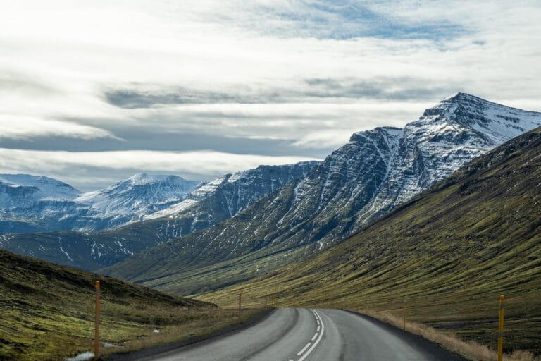 A winding Iceland road leads towards majestic snow-capped mountains under a partly cloudy sky. The scene evokes a sense of adventure and tranquility.