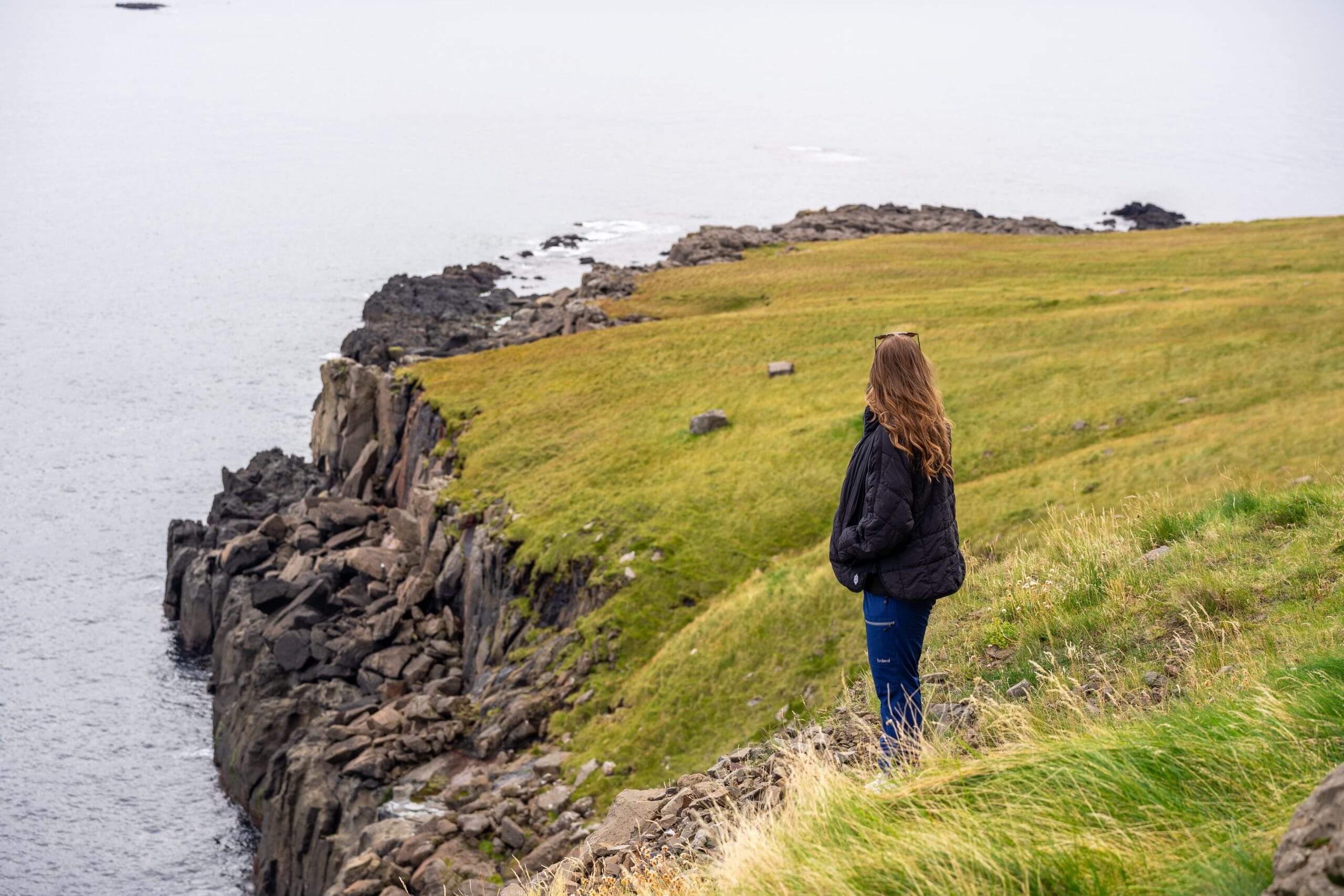 A person with long hair stands on a grassy cliff edge overlooking the ocean. The rocky coastline creates a serene and contemplative atmosphere.