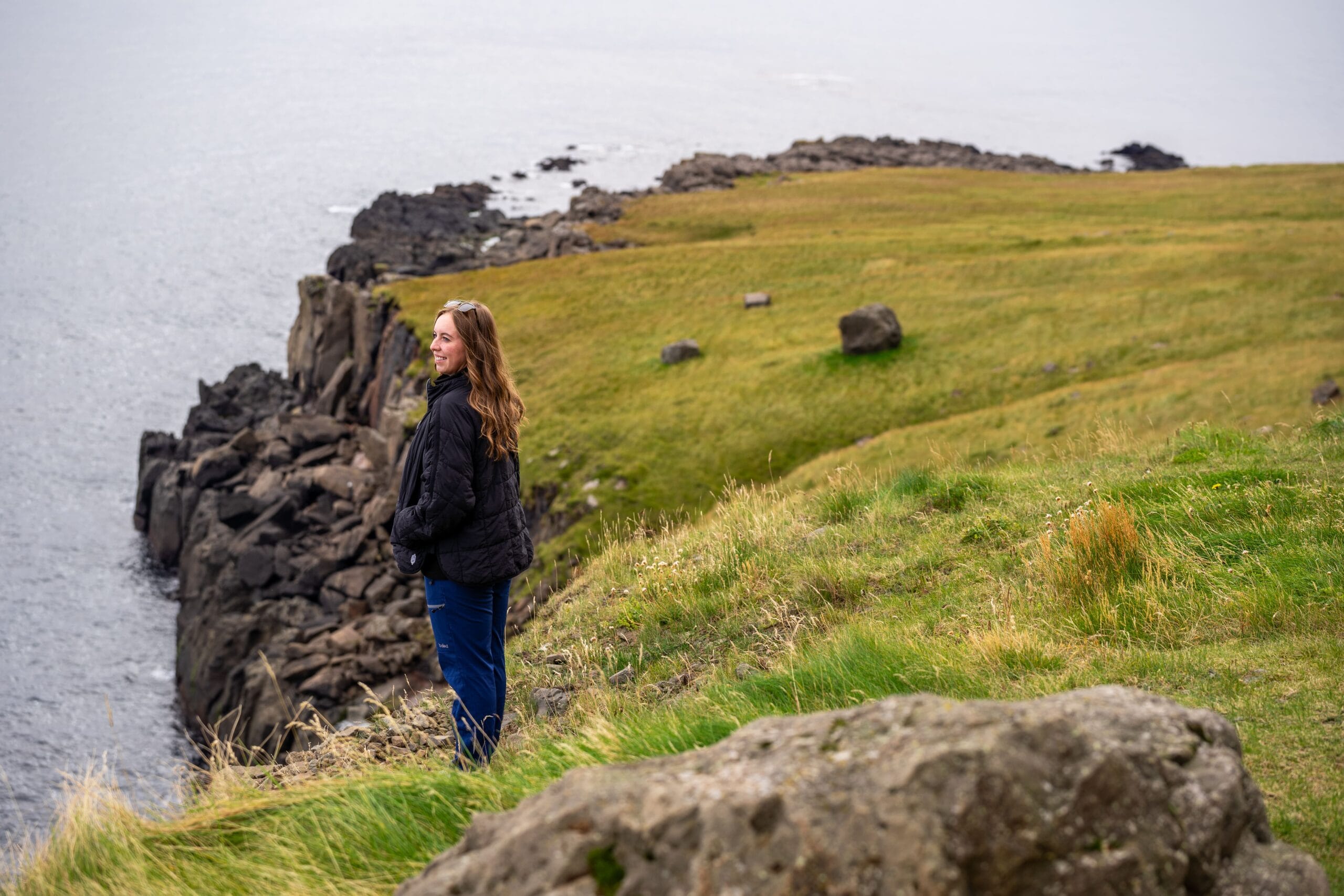 A woman in a black jacket and blue pants stands on a grassy cliff, looking toward the rocky coastline and the sea beyond, evoking a sense of calm and freedom during an iceland road trip.