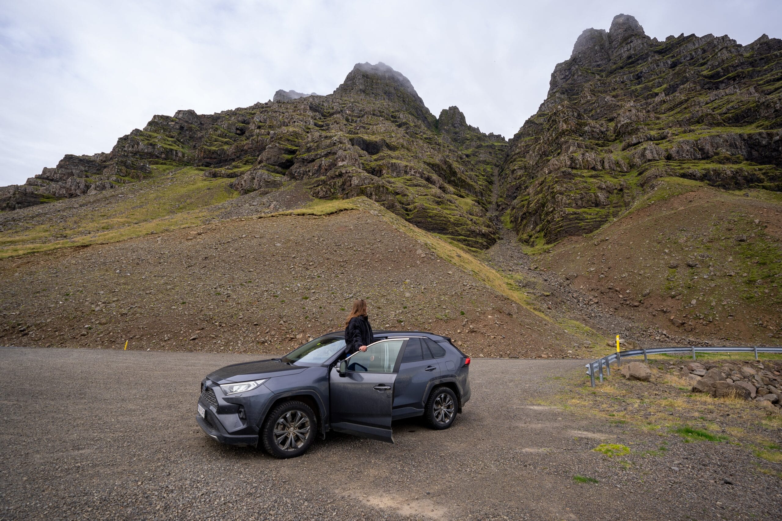 A person stands beside an open car door, facing rugged, rocky mountains under a cloudy sky, conveying a sense of adventure and exploration.