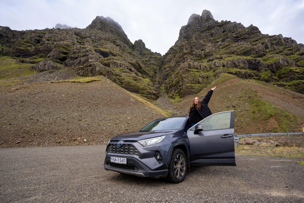 A smiling person stands beside an open car 4x4 rental in iceland, pointing upward amidst dramatic, rocky hills under a cloudy sky, conveying excitement and adventure.