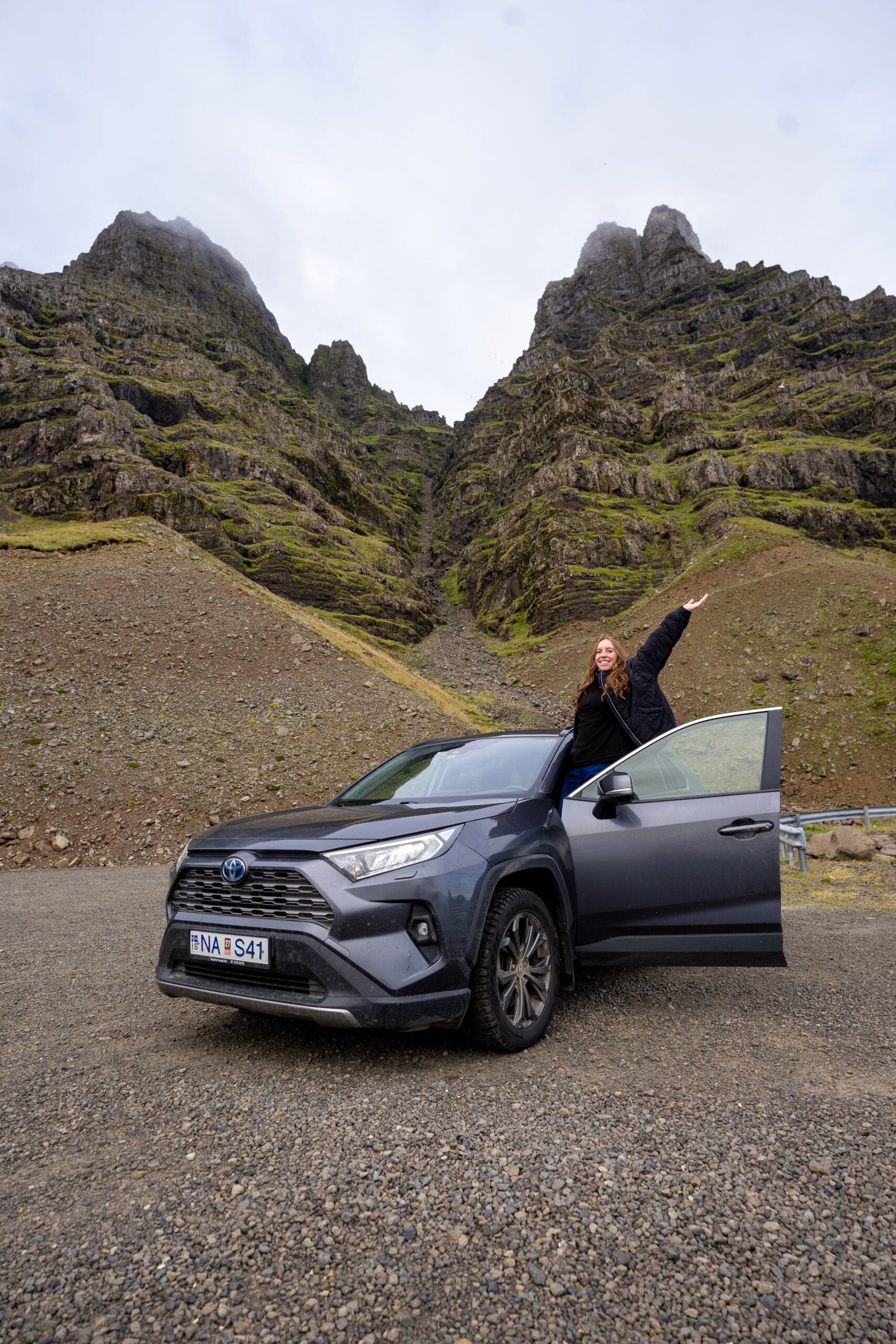 A person stands beside an open car door on a rocky terrain, pointing toward towering, moss-covered mountains under a cloudy sky. The mood is adventurous.