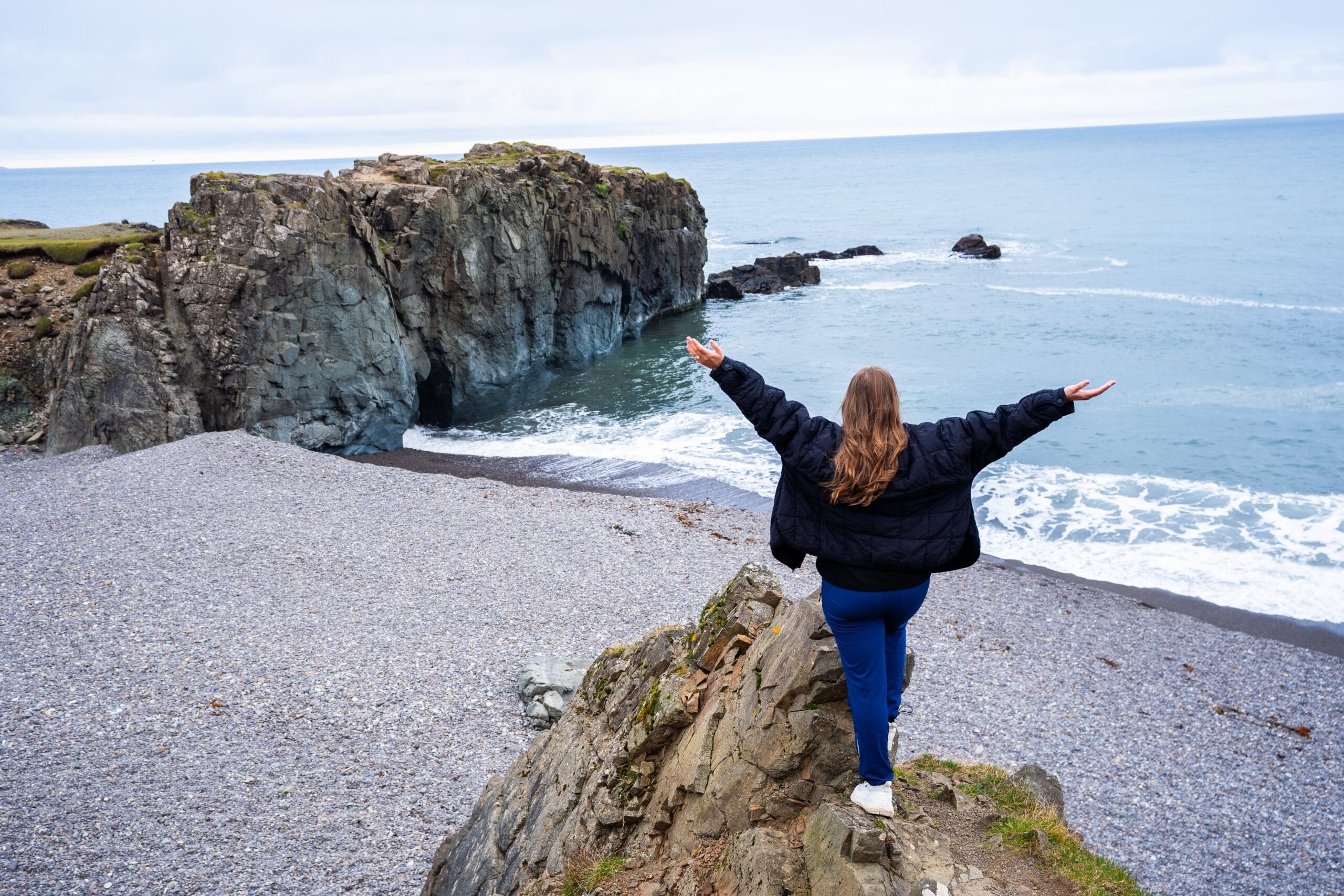 Person stands on a rocky cliff edge with arms outstretched, facing a vast ocean. Overcast sky and waves hitting the pebble beach convey serenity.