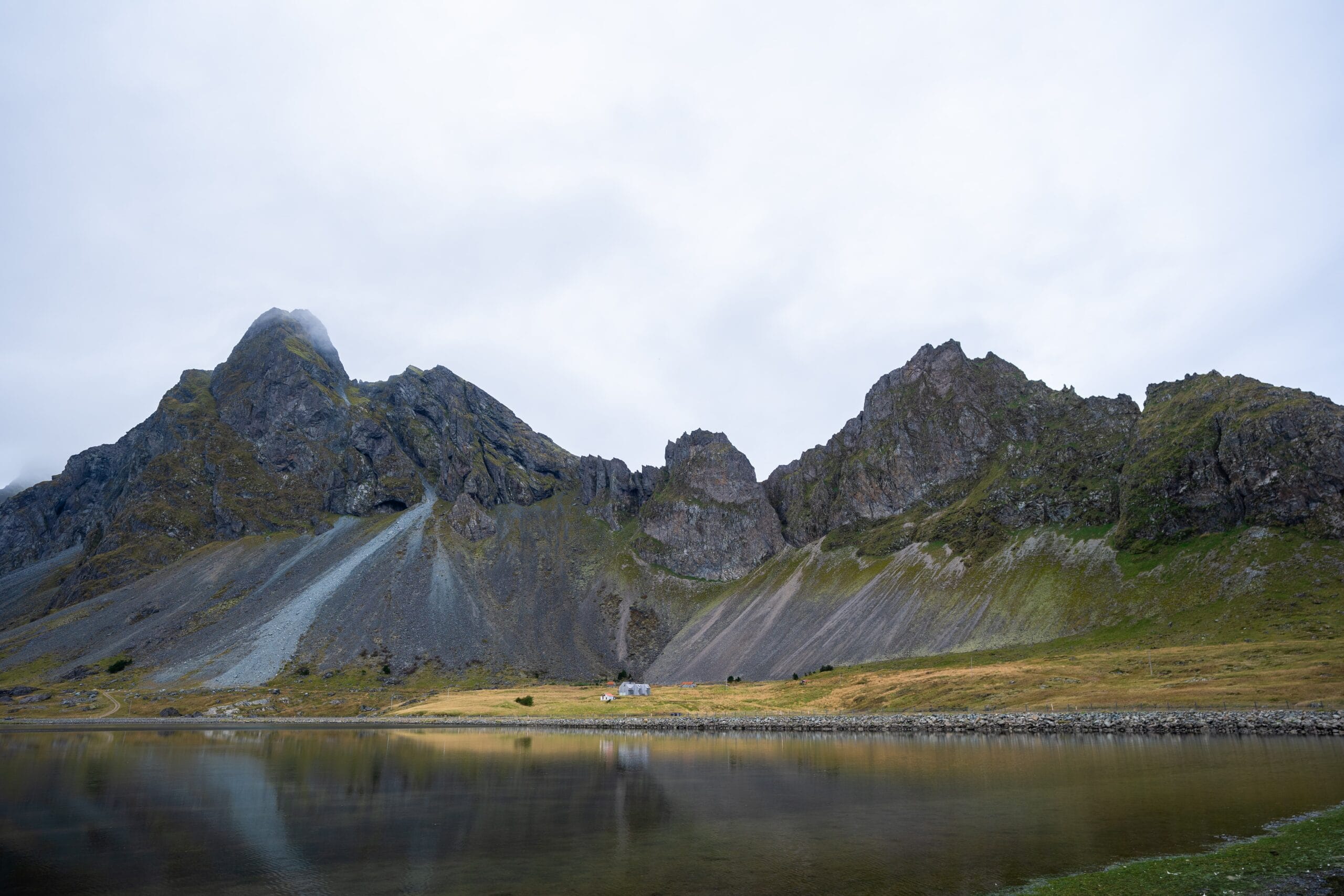 A rugged mountain range towers over a calm lake, with a small building at the base. The overcast sky adds a serene yet dramatic tone to the scene.