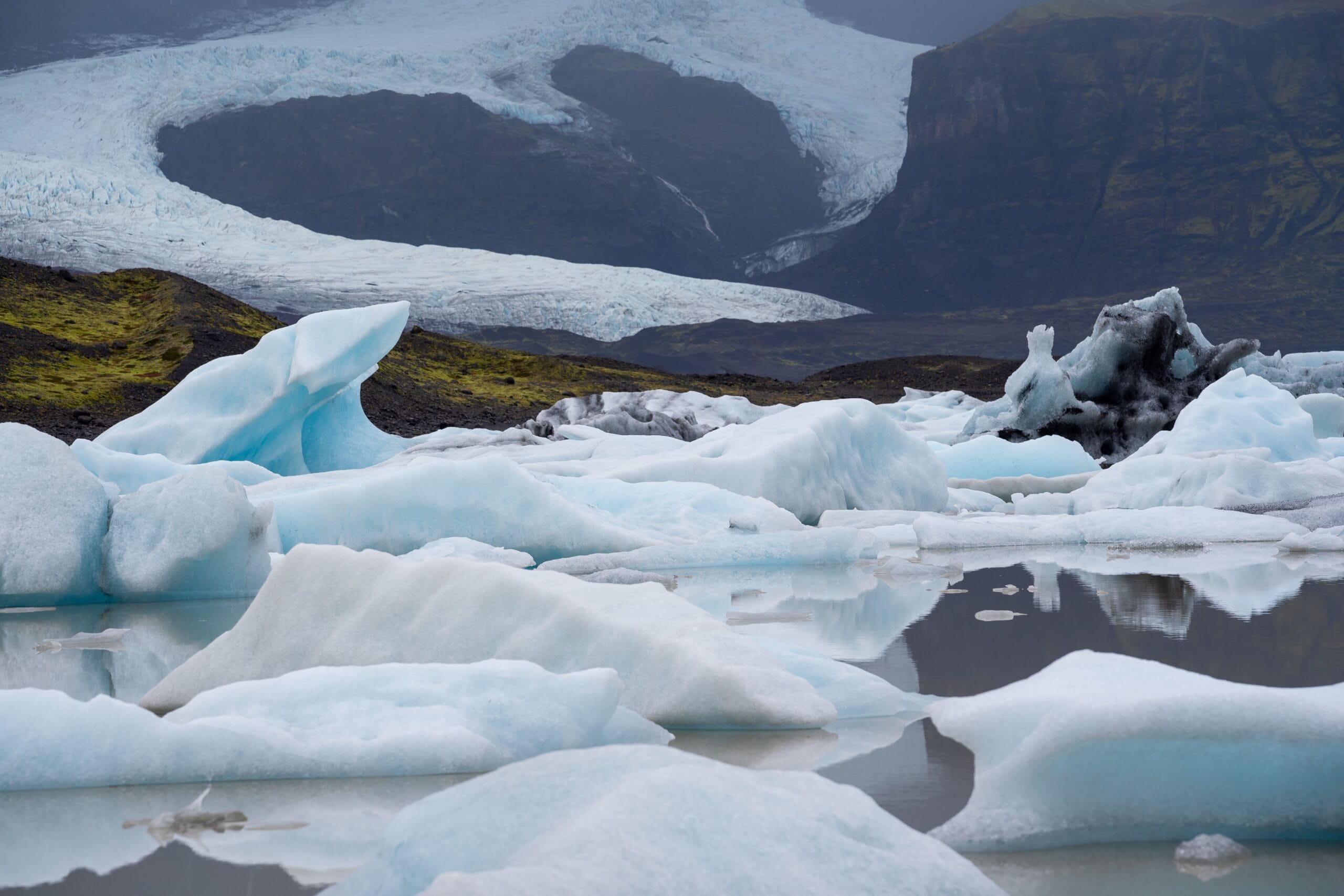 Icy landscape with scattered blue icebergs floating on a calm lake. In the background, a winding glacier stretches into misty, cloudy mountains.