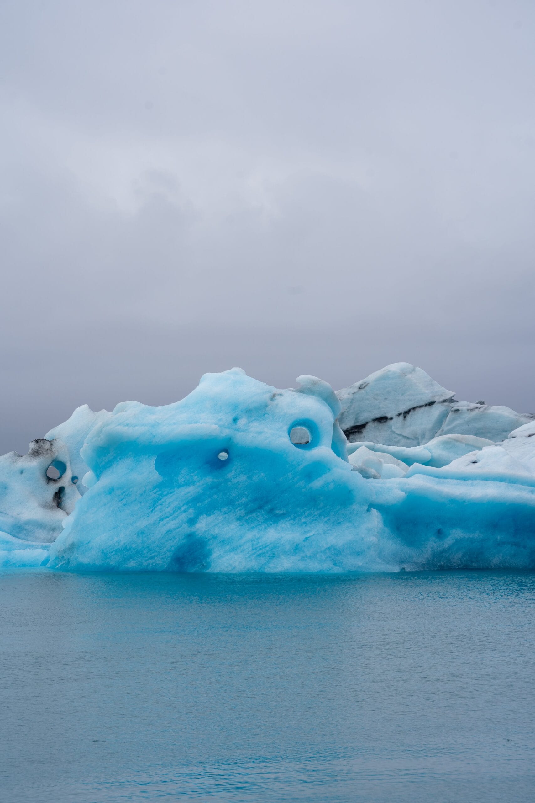 A large, jagged iceberg with deep blue hues floats in calm, turquoise water under an overcast sky, conveying a serene and chilly atmosphere.