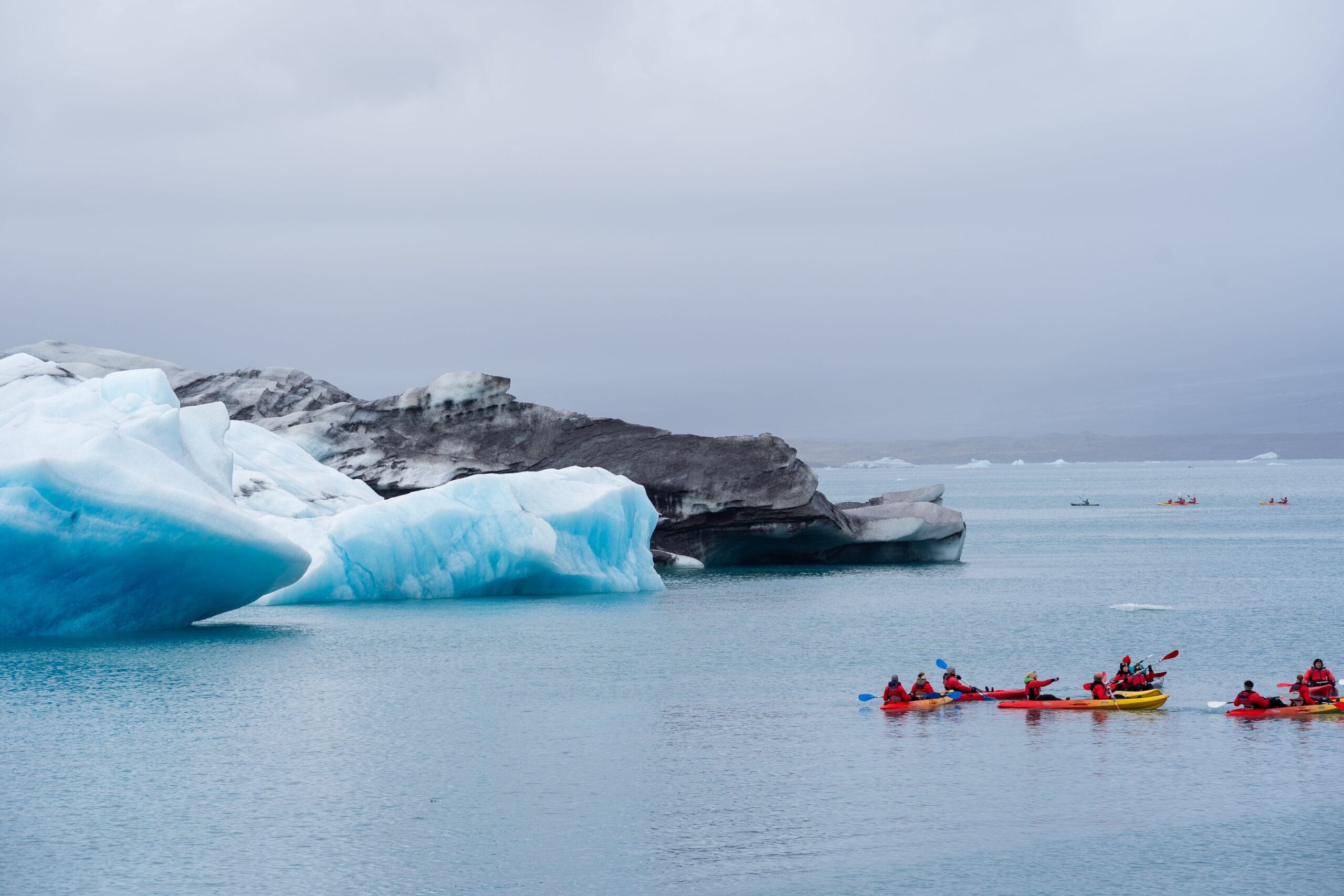 Kayakers in red jackets navigate icy blue waters near a large glacier under a cloudy sky, conveying adventure and tranquility in a cold landscape.