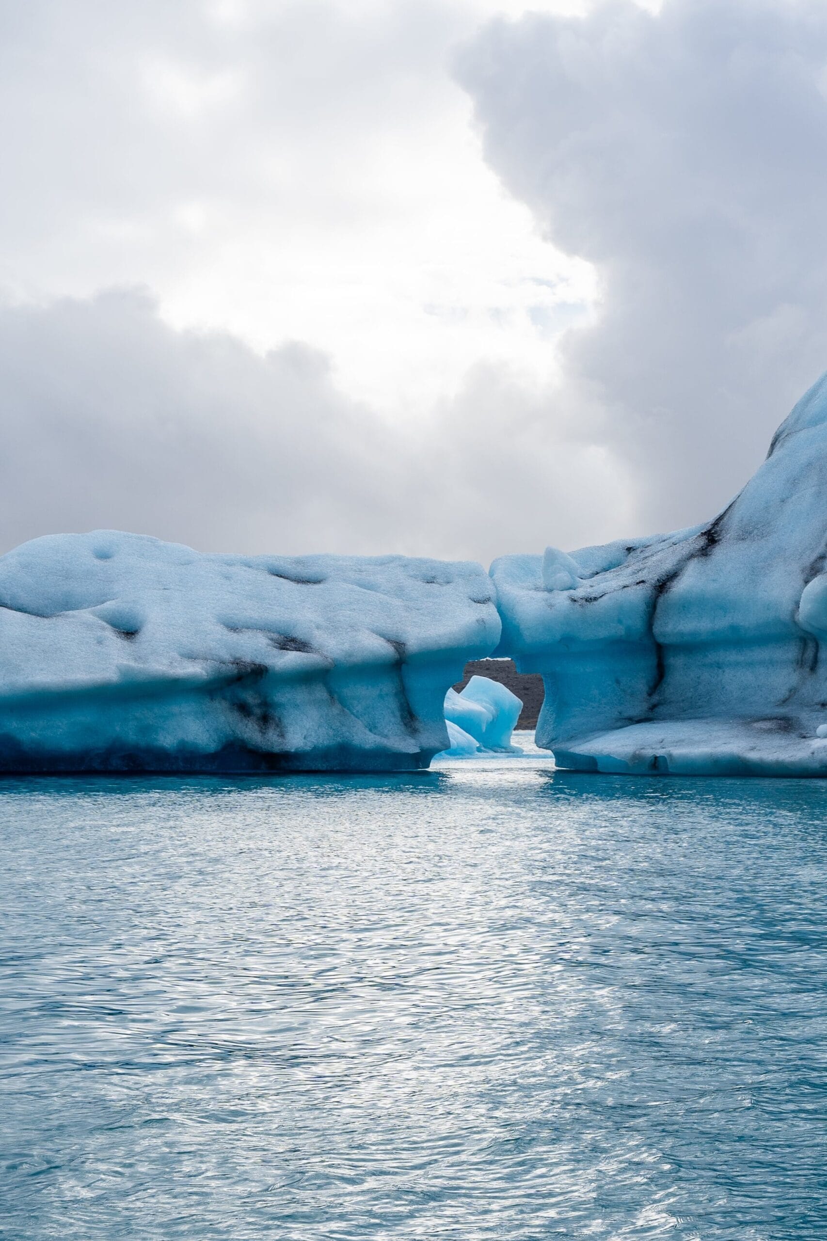 A large blue iceberg floats in calm sea waters under a cloudy sky. The iceberg has a distinctive arch shape, with a hole near the base. The scene is peaceful and majestic.