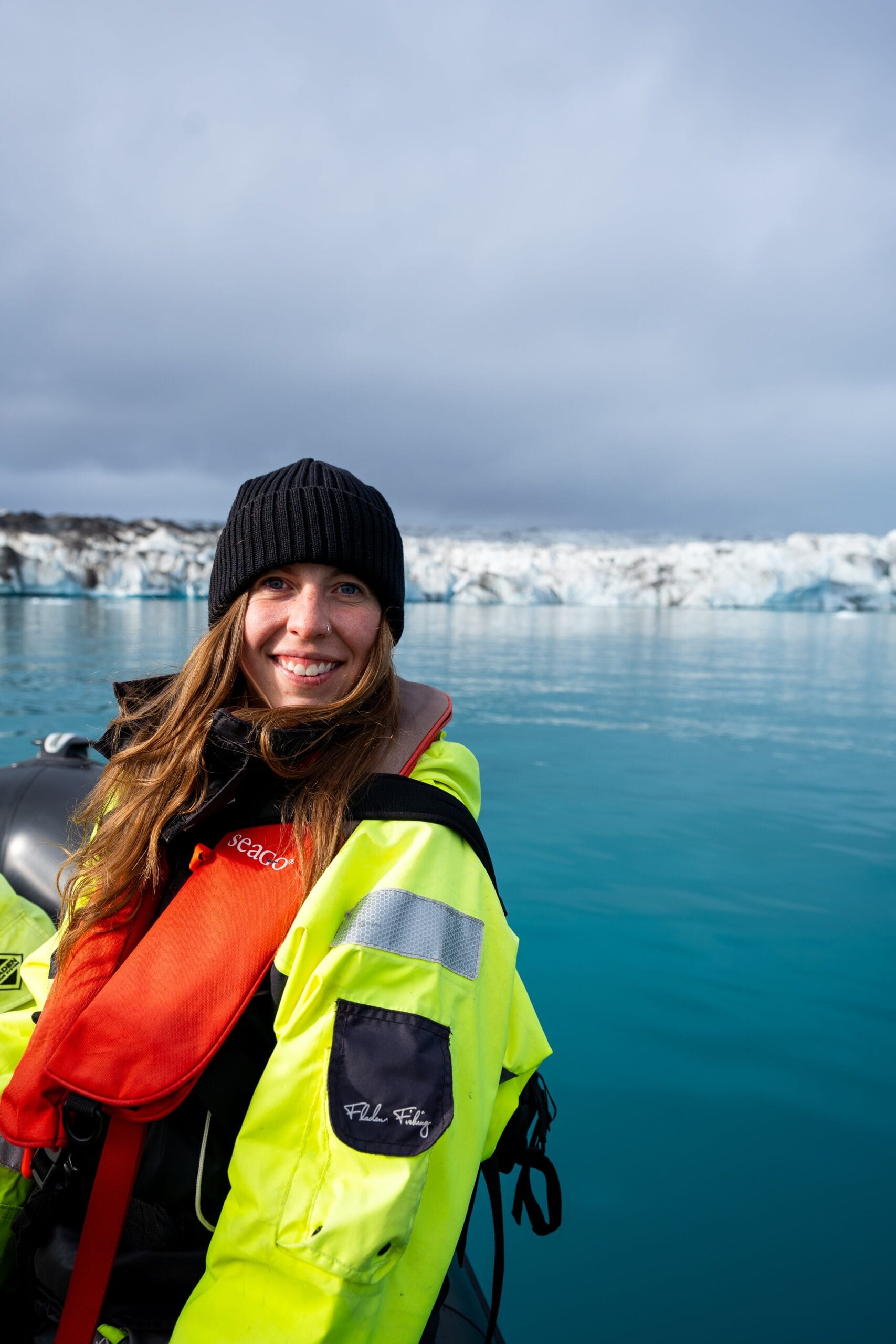 Woman on Iceland excursions in a bright yellow jacket and black beanie smiles on a boat in icy blue water, with a glacier in the background under a cloudy sky.