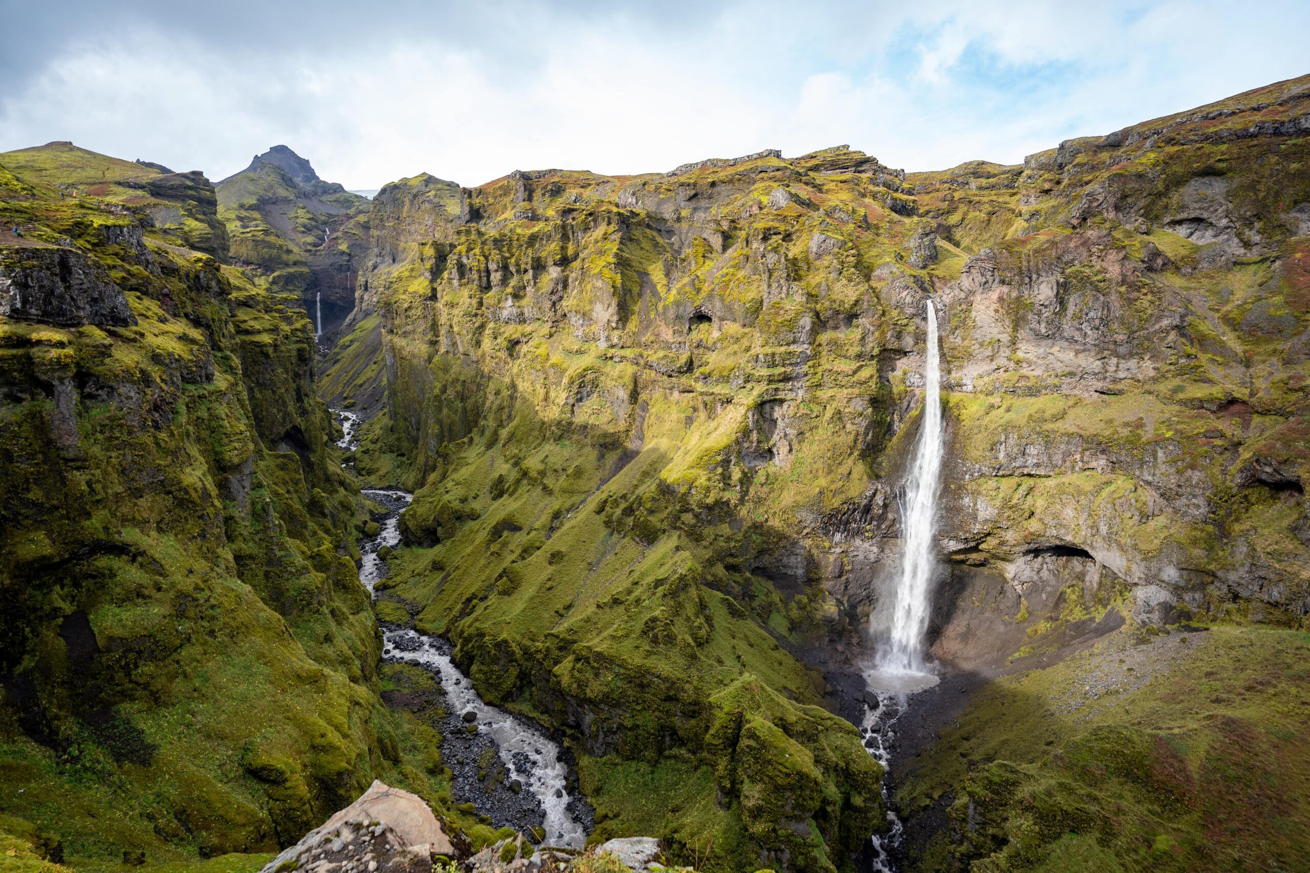 A tall, narrow waterfall cascades down a lush, green cliff into a winding river below, set against a backdrop of rugged mountains and a bright sky.