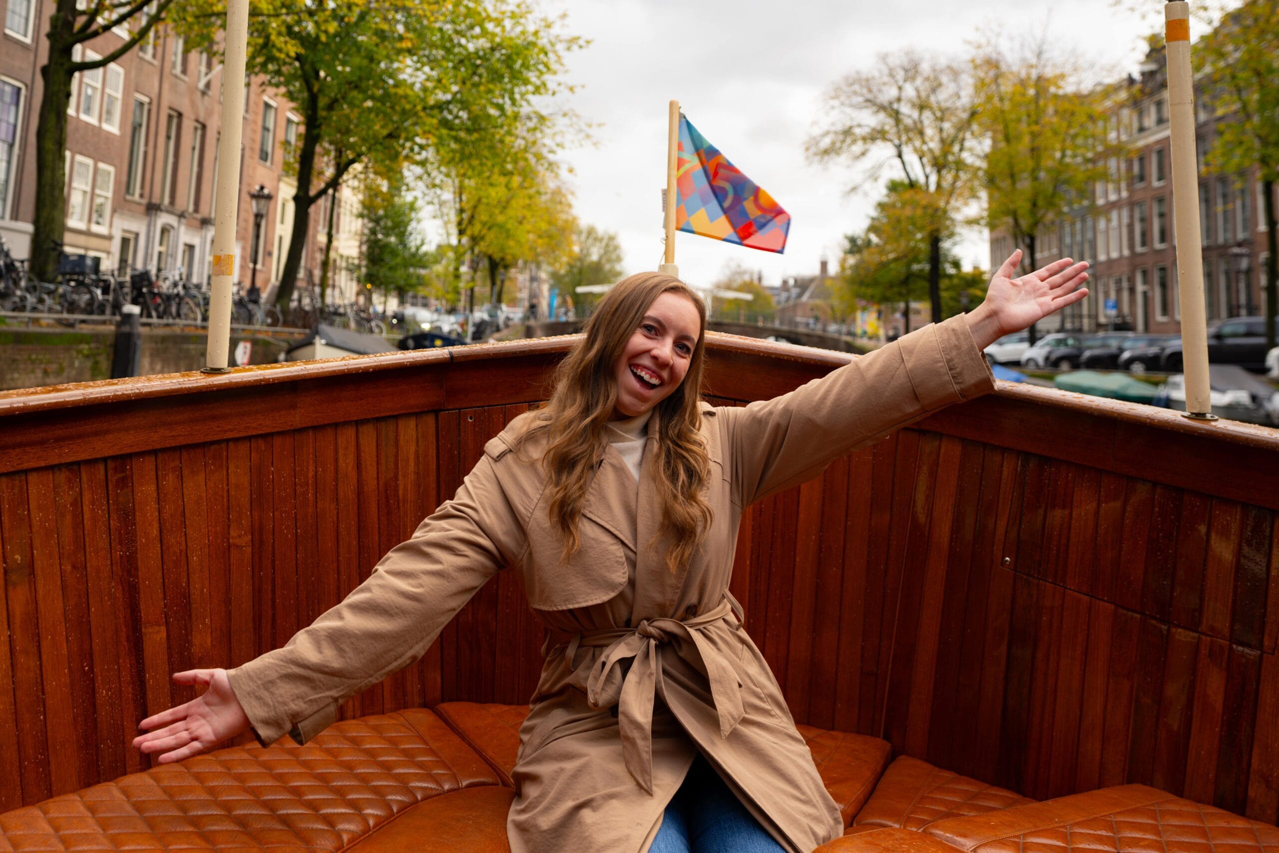 A woman in a beige coat joyfully poses with arms outstretched on a wooden boat in a canal, vibrant flag above, surrounded by trees and buildings.