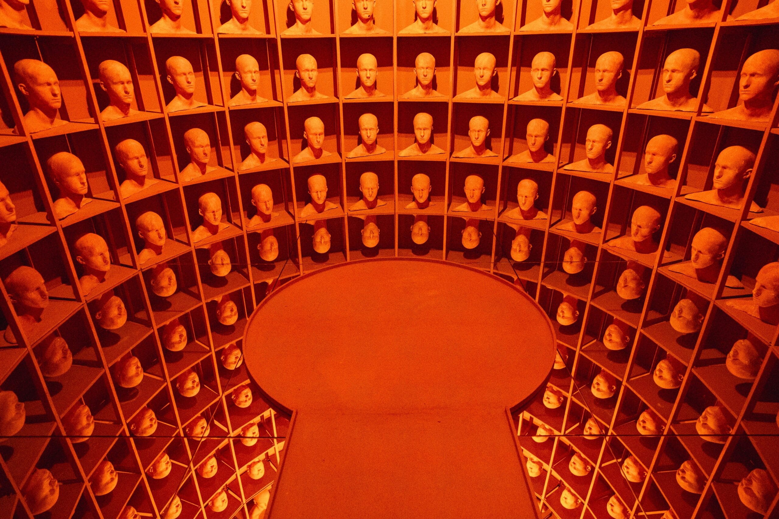 Room with walls of identical mannequin heads in orange lighting, creating an eerie and unsettling atmosphere. Floor mirrors enhance the infinite pattern.
