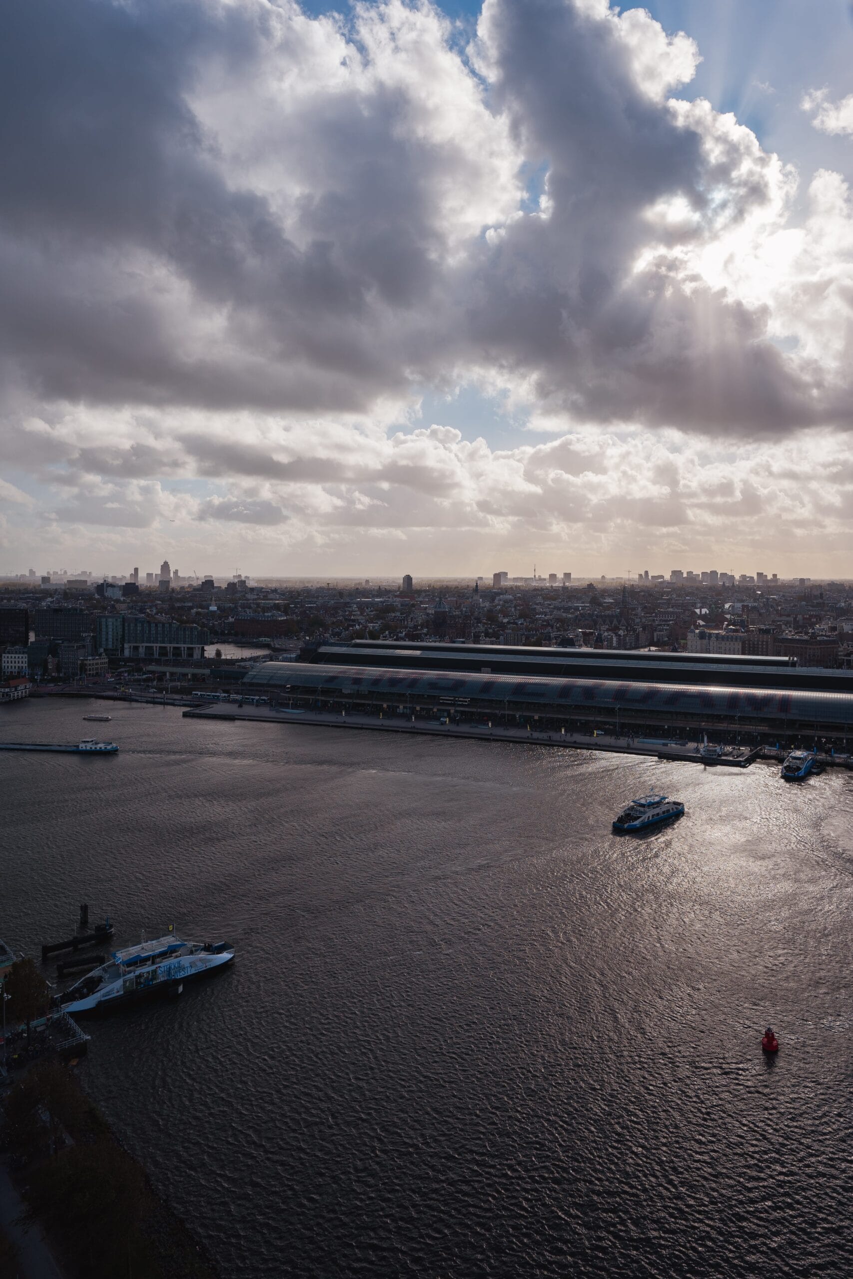 A vast Amsterdam river reflects sunlight under a dramatic sky with sunbeams piercing thick clouds. A city skyline and large building silhouette the horizon.