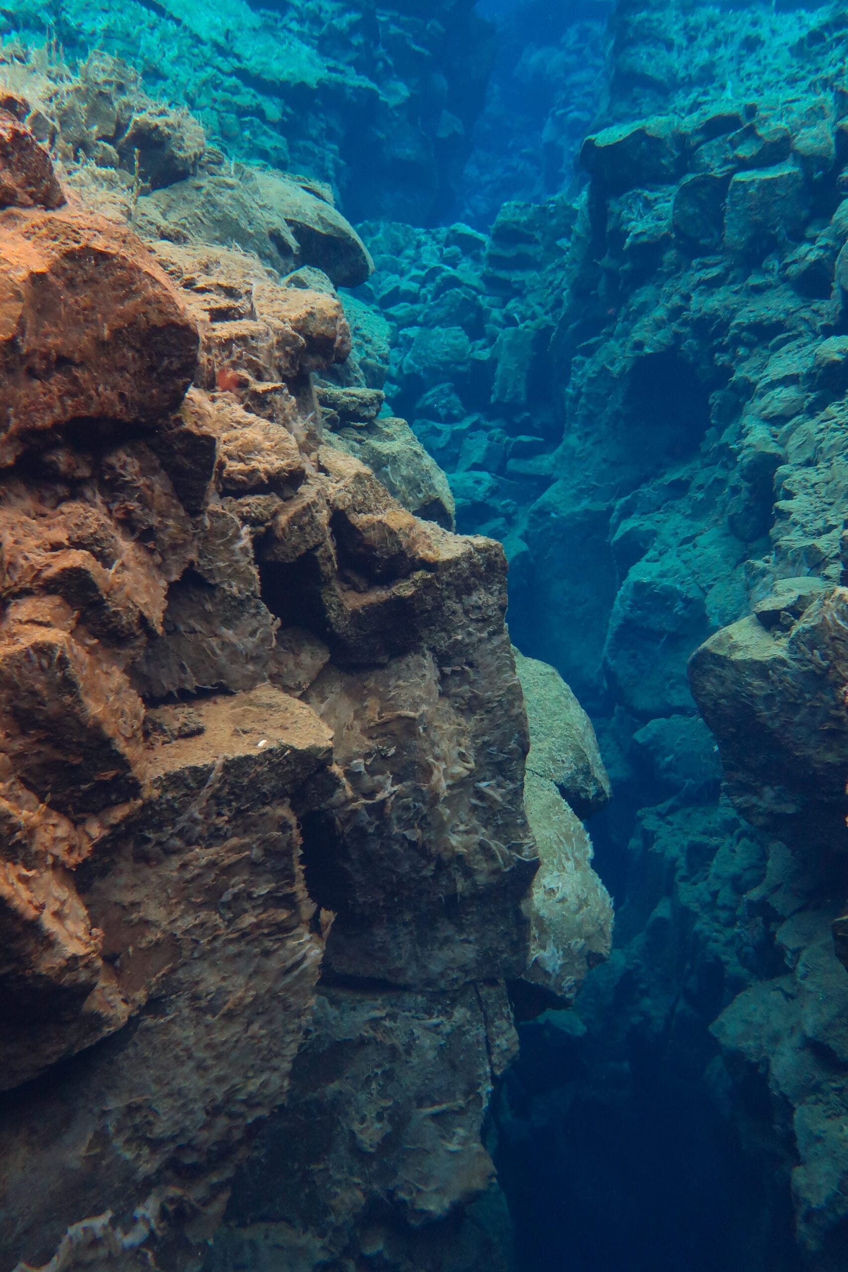 Underwater view of a deep, narrow ravine with jagged rocky walls. Clear blue water reveals textured rocks, evoking a sense of mystery and exploration.