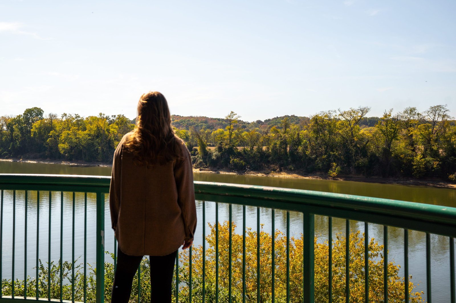 Woman in a brown jacket looking out over the Cumberland River from a green railing on the Clarksville Riverwalk in fall